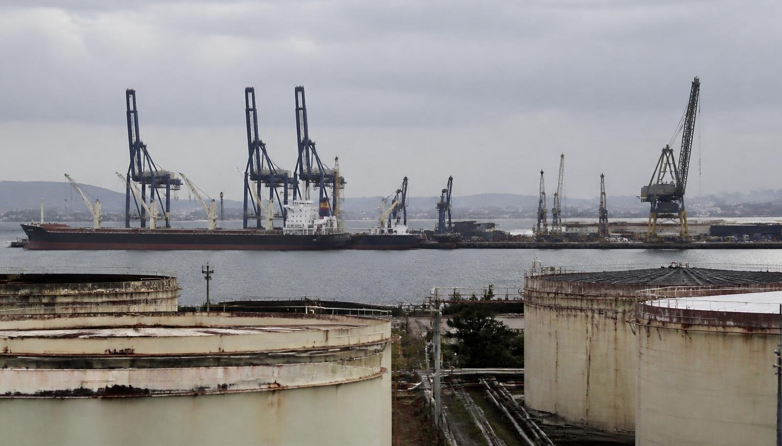 Terminal portuário industrial na Bahia com guindastes, navio atracado e tanques de armazenamento em primeiro plano sob céu nublado
