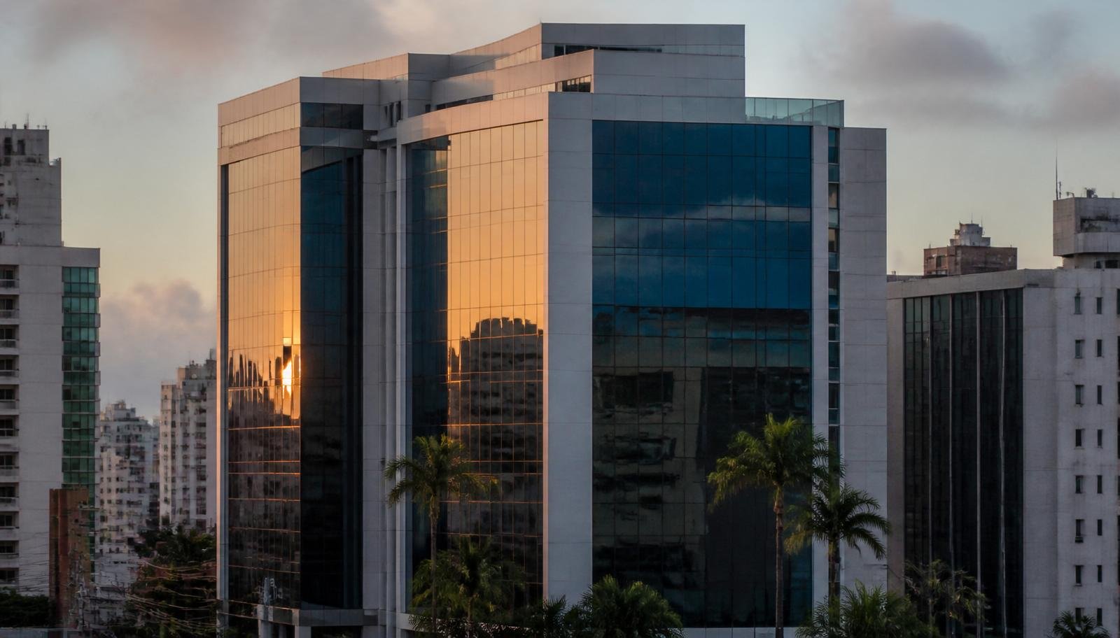 Edifício comercial corporativo em Salvador, Bahia, ao entardecer, com fachada envidraçada refletindo o pôr do sol e coqueiros na frente, típico da região onde funcionam escritórios de advocacia, contabilidade e planejamento sucessório especializados em holdings patrimoniais