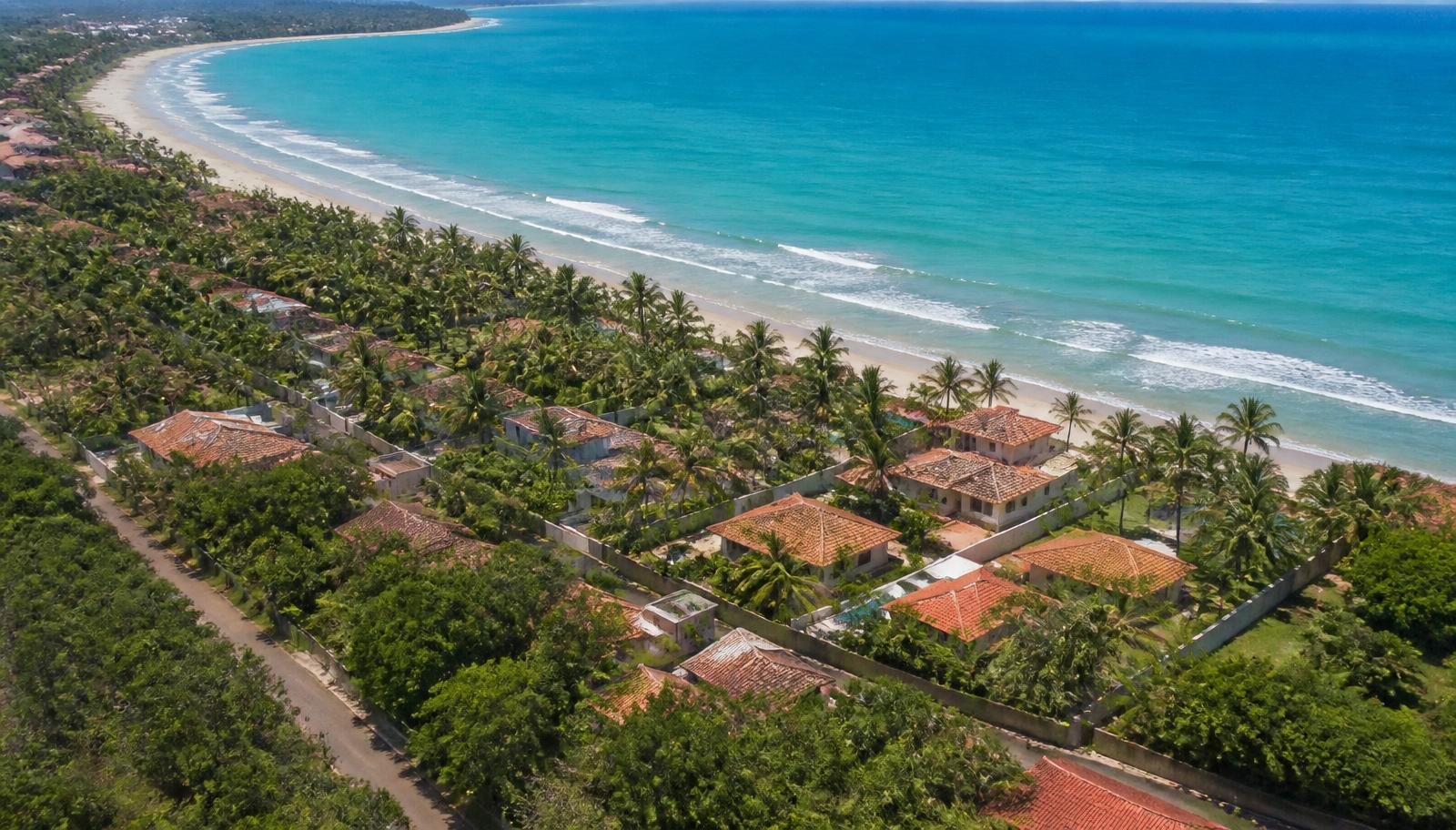 Vista aérea de casas e pousadas de temporada distribuídas ao longo da orla no litoral sul da Bahia, com mar turquesa, coqueiros e telhados de terracota